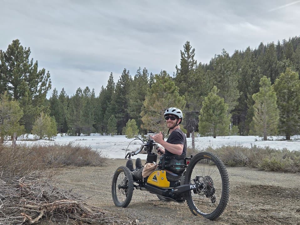 Caleb grinning on an off-road adaptive handcycle in a snowy pine forest